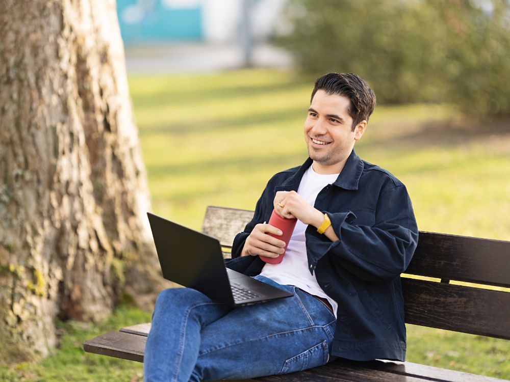 Um funcionário da Henkel se senta em um banco de um parque e trabalha em seu laptop. 