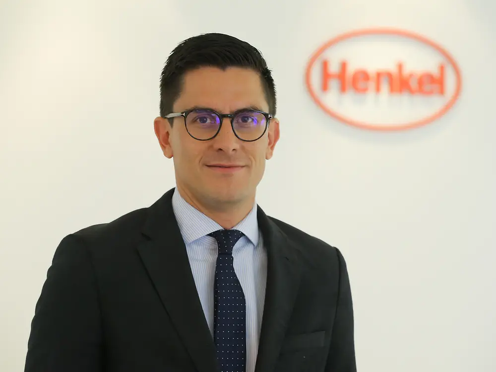 André Barón – André em pé, usando um terno preto. Ao fundo, parede branca com o logo da Henkel. 