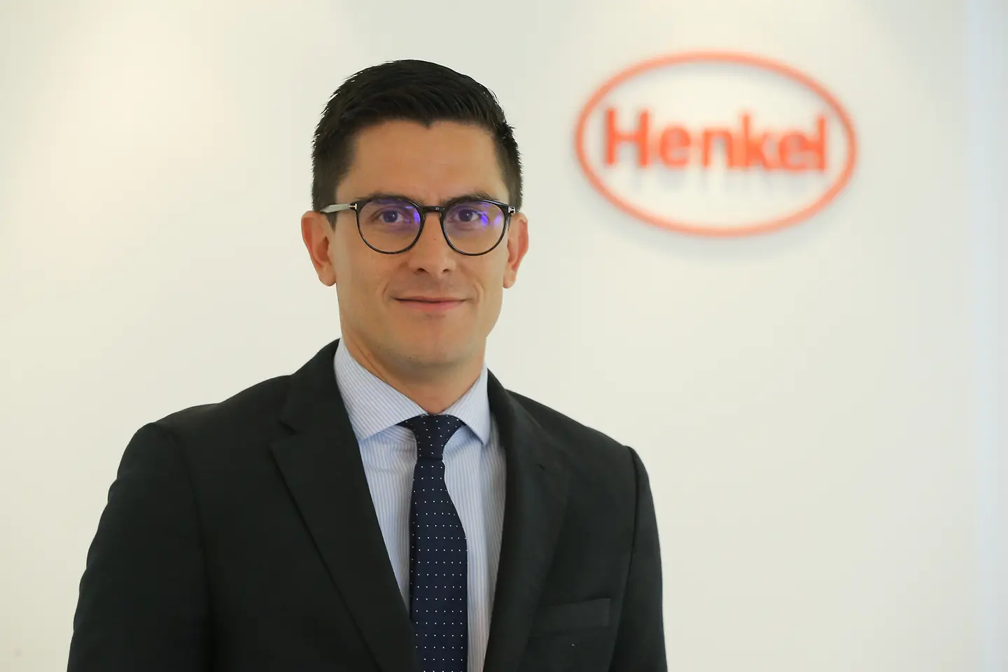 André Barón – André em pé, usando um terno preto. Ao fundo, parede branca com o logo da Henkel. 