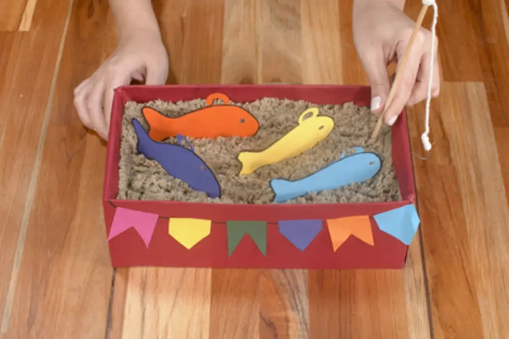 Caixa vermelha com areia dentro e peixes coloridos de papel. Na frente da caixa, bandeiras coloridas.