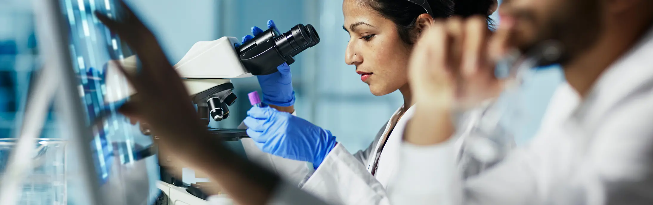 um laboratório com uma mulher sentada em frente a um microscópio, olhando uma amostra e na frente um homem com barba olhando para uma tela 
