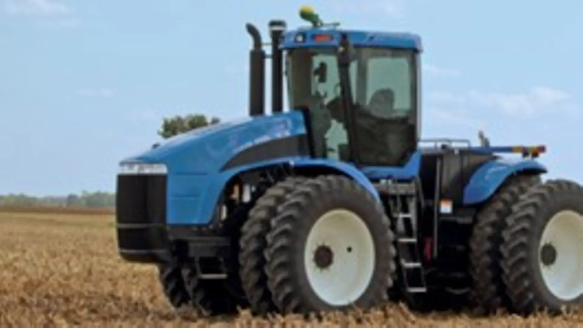 Trator Agrícola – Trator azul sobre o campo com folhagens amareladas. Ao fundo, paisagem montanhosa.
