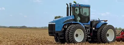 Trator Agrícola – Trator azul sobre o campo com folhagens amareladas. Ao fundo, paisagem montanhosa.