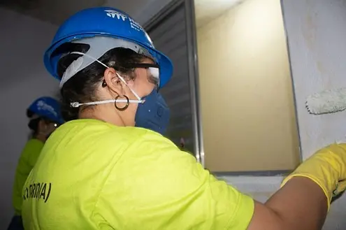 mulher com capacete, máscara e luva de proteção pintando uma parede perto da janela