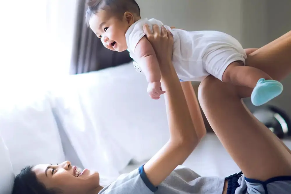 Mãe levanta e brinca com um bebê sorridente dentro de casa, em um ambiente iluminado.