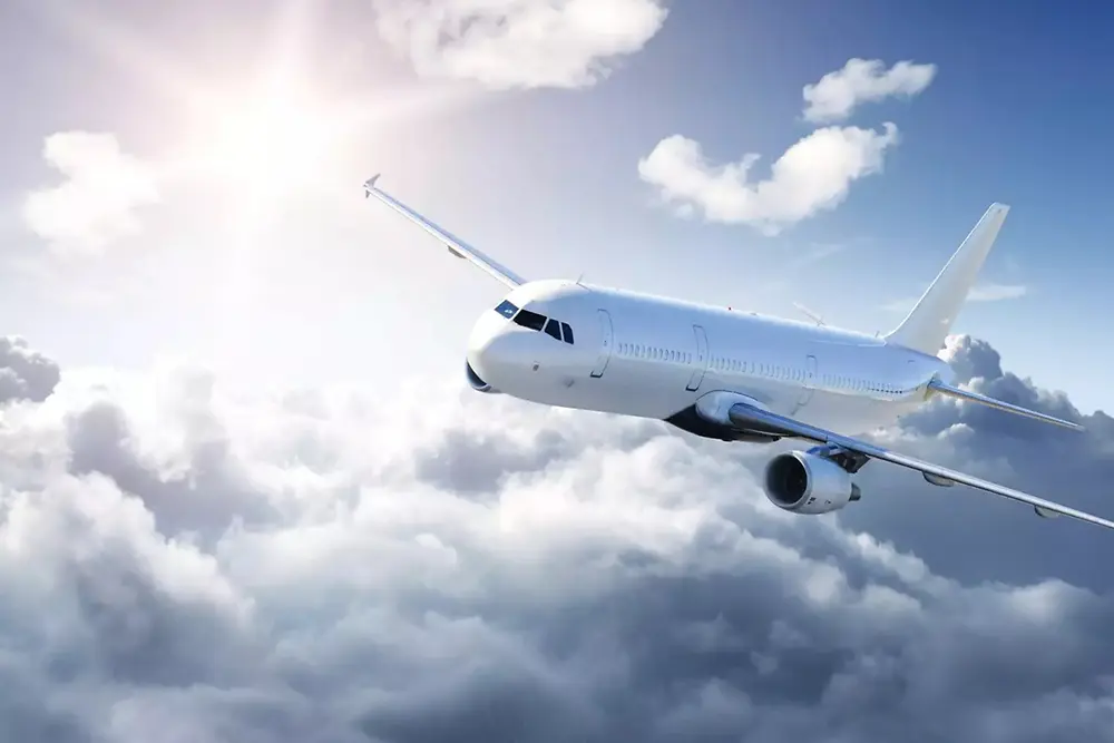 Um avião de passageiros branco voando acima de nuvens suaves em um céu claro, com a luz do sol brilhando no canto superior esquerdo.