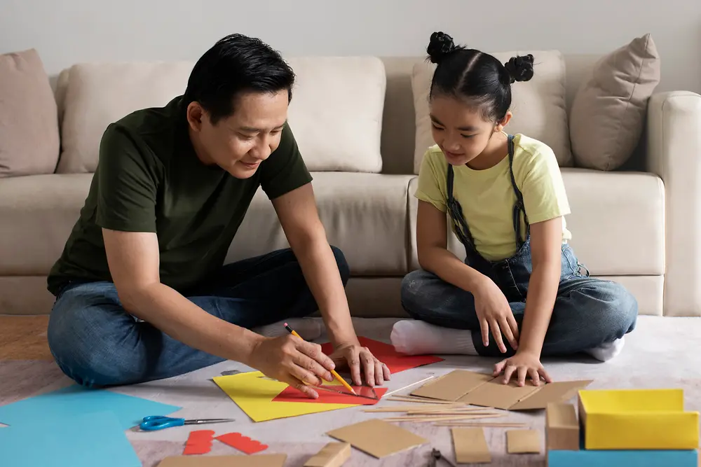 Pai e filha se unem para exercitar a criatividade em trabalhos manuais com o uso de papeis, cola e outros materiais.