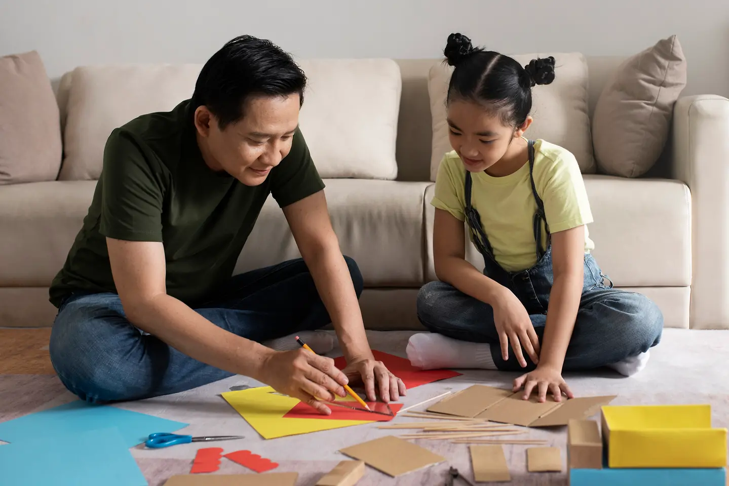 Pai e filha se unem para exercitar a criatividade em trabalhos manuais com o uso de papeis, cola e outros materiais.