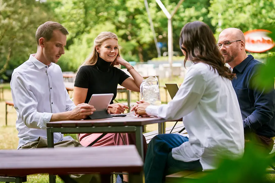 Grupo de colegas sentados juntos ao ar livre, discutindo trabalho com tablets e notebooks, representando a colaboração e uma cultura aberta e orientada para a equipe na Henkel.