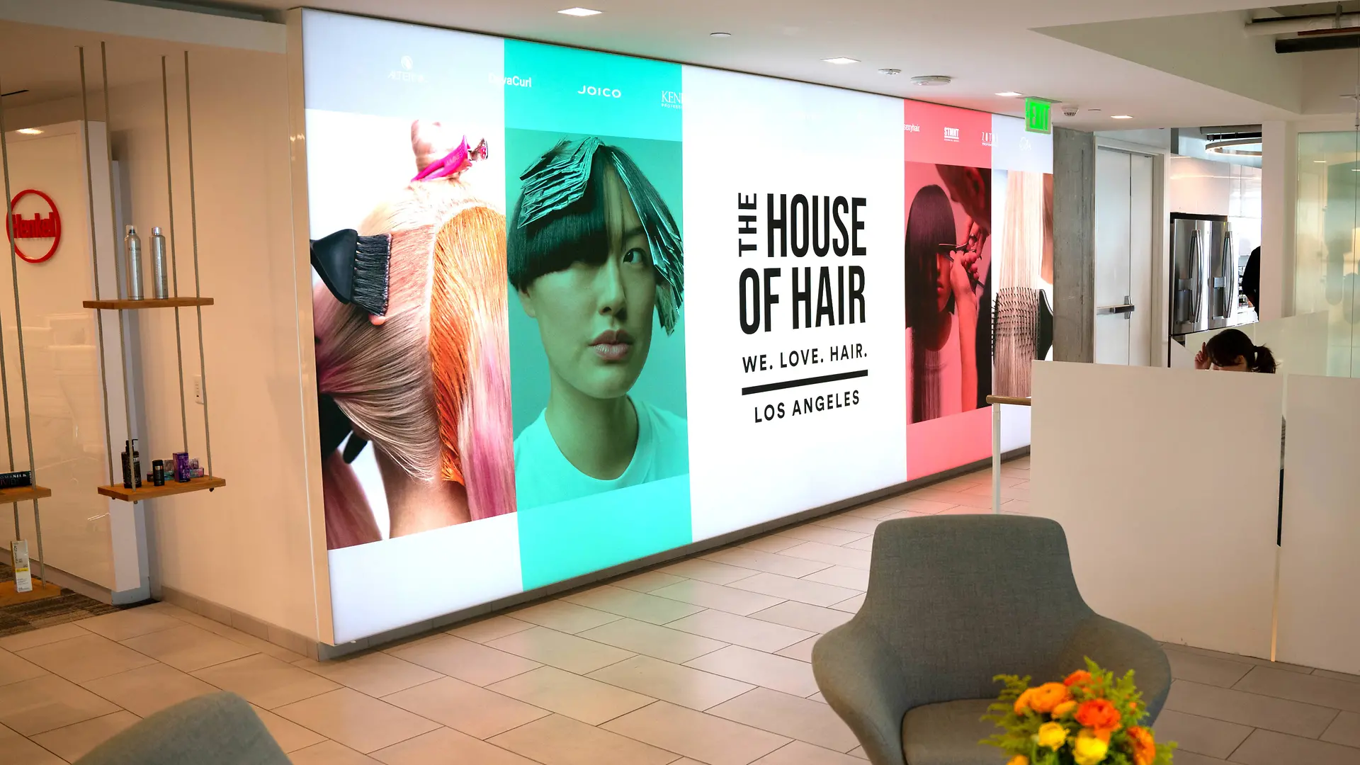 Lobby moderno com um painel iluminado exibindo grandes gráficos coloridos com tema de cabelo e o texto “THE HOUSE OF HAIR – WE. LOVE. HAIR. – LOS ANGELES.” O espaço inclui poltronas cinza-claro, uma mesa com arranjo floral vibrante e design limpo e minimalista com piso revestido e divisórias brancas.