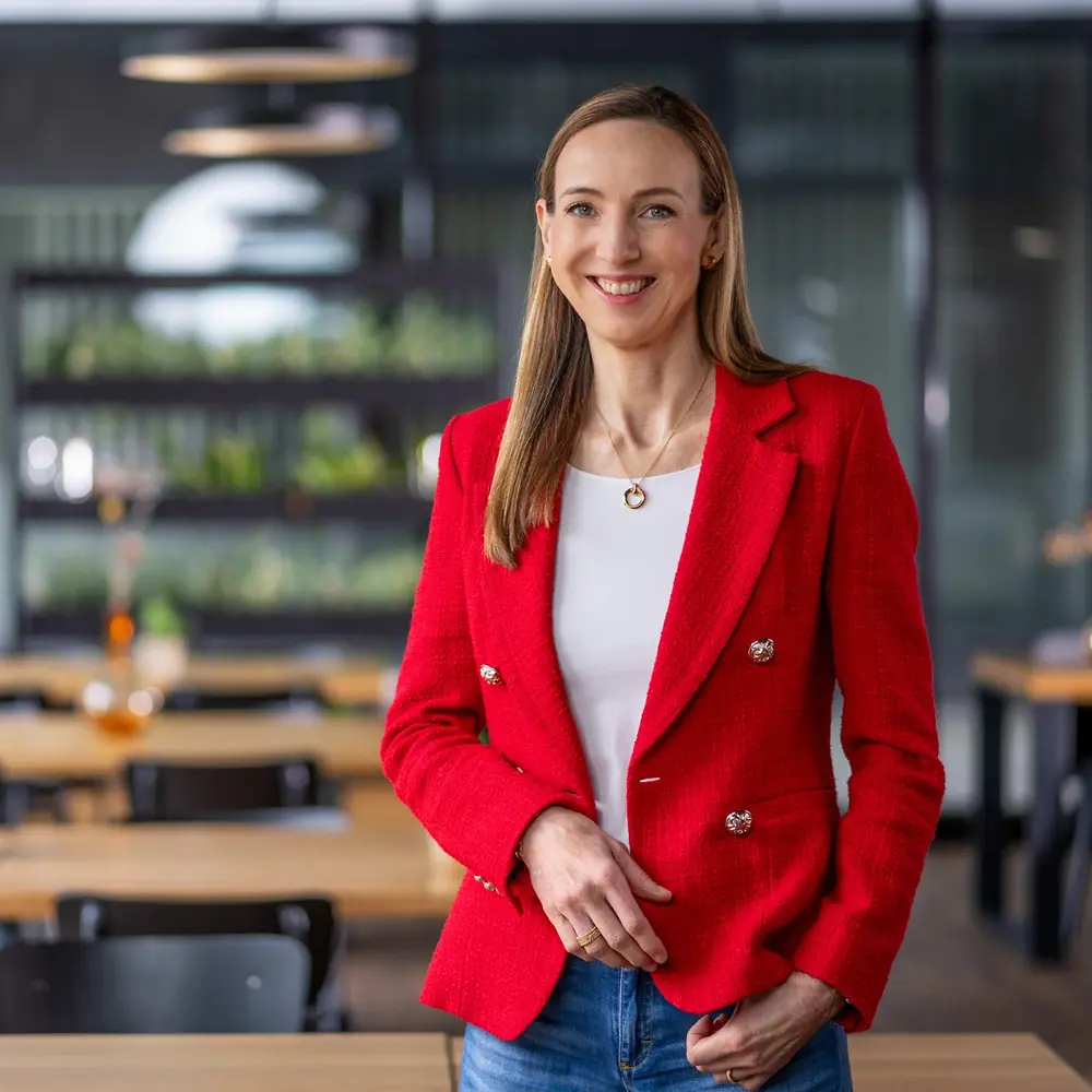 Dra. Simone Bagel-Trah vestindo um blazer vermelho vivo e uma blusa branca, em pé em um ambiente moderno estilo café, com mesas de madeira e vasos decorativos ao fundo.