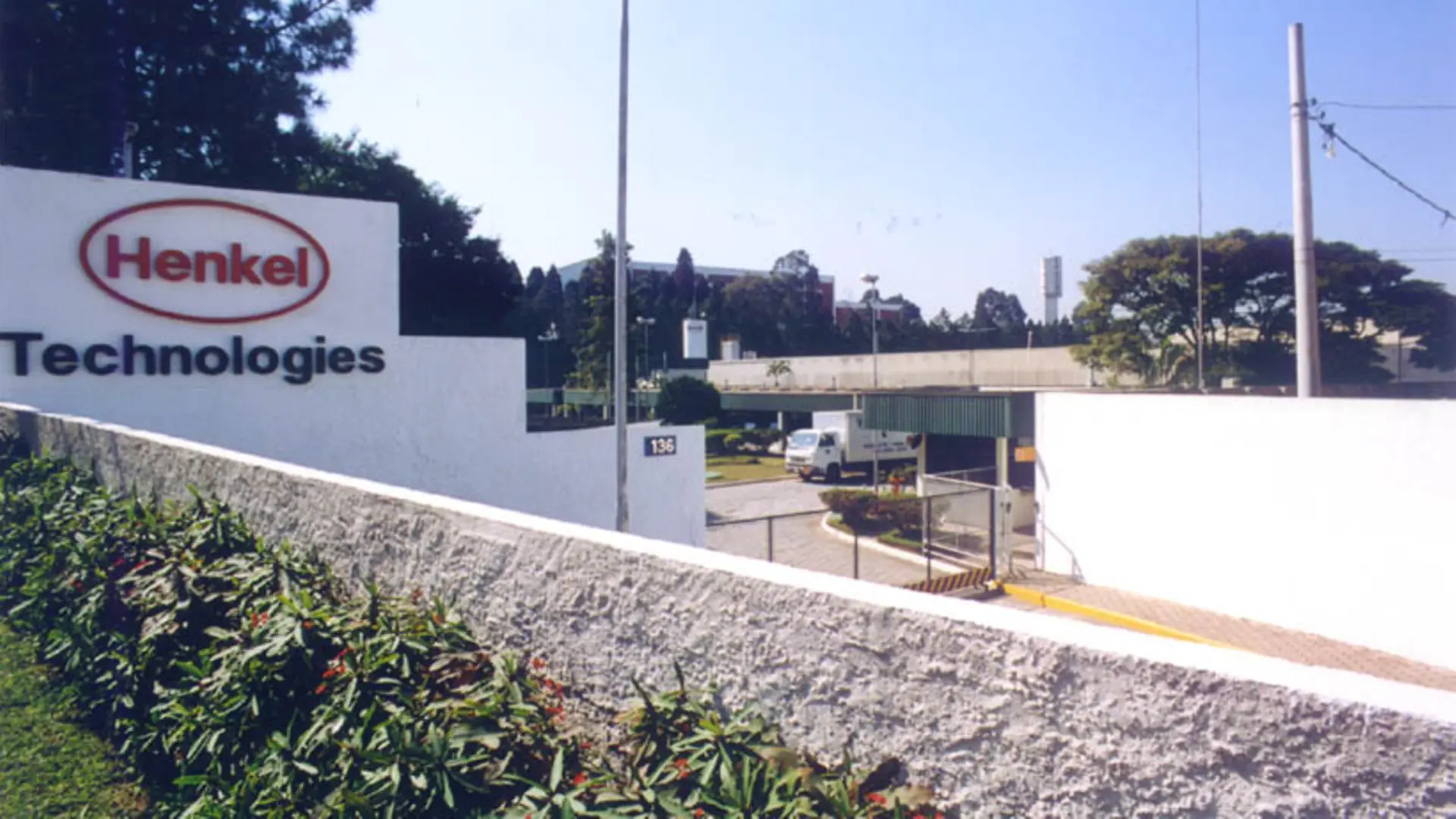 Entrada da unidade Henkel Technologies, em Diadema, com muro branco exibindo o logotipo e o nome da empresa. Em primeiro plano, há vegetação ornamental; ao fundo, portaria, guarita de segurança e prédios industriais sob céu azul