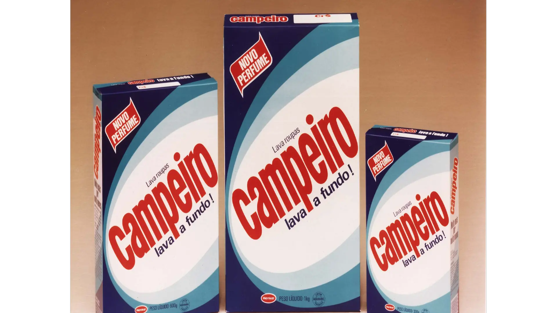 Três caixas do detergente em pó Campeiro, com design em tons de azul e vermelho, exibindo o texto ‘Novo Perfume’ e o slogan ‘Lava roupas Campeiro lava a fundo!’.