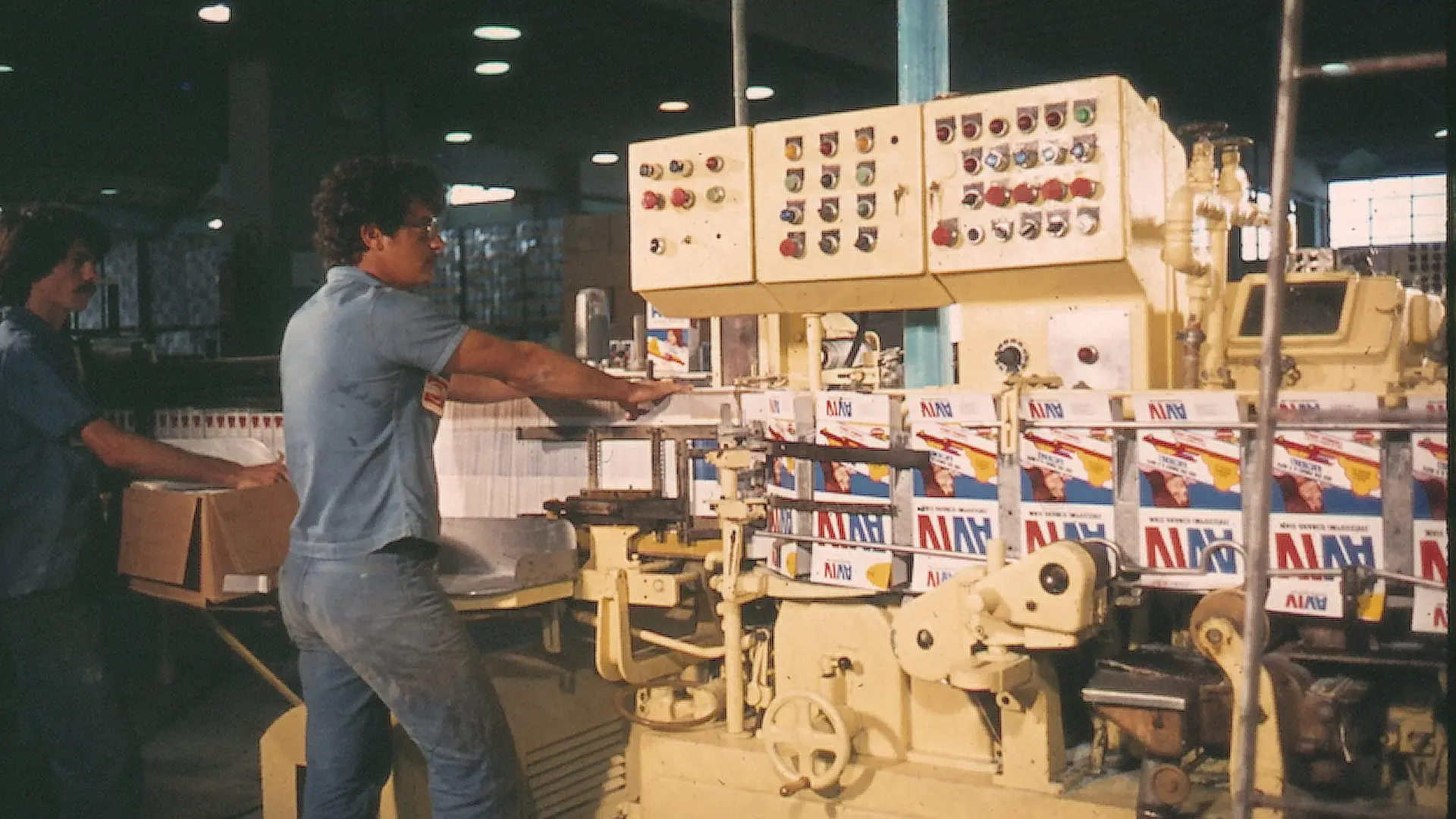 Linha de produção industrial com máquina embaladora e caixas do detergente Viva sendo processadas, em ambiente fabril com iluminação artificial e painéis de controle visíveis.