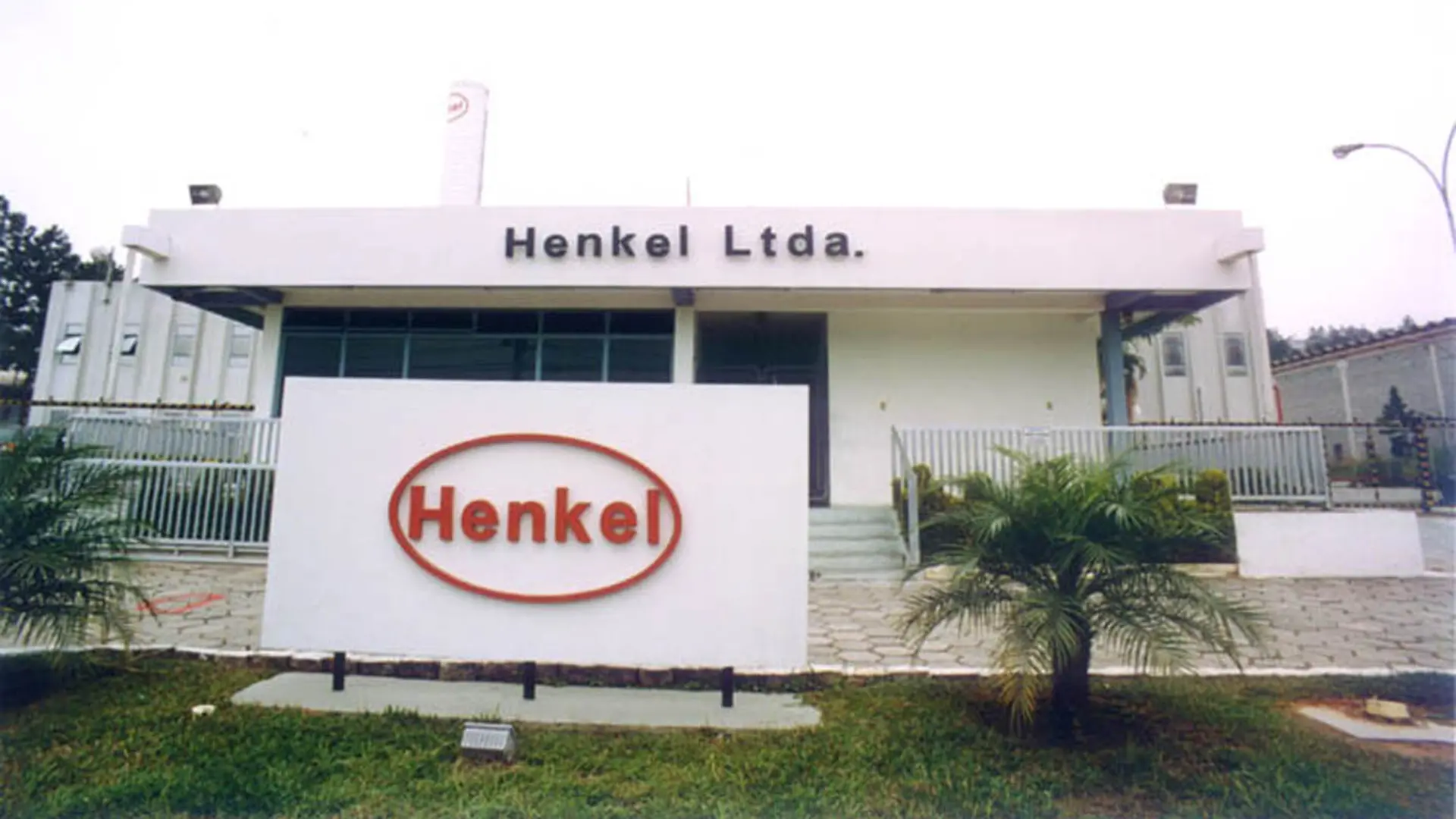 Fachada da unidade Henkel Ltda. em Itapevi, com um prédio branco de linhas retas, letreiro 'Henkel Ltda.' no topo e um grande painel frontal com o logotipo oval vermelho da Henkel.