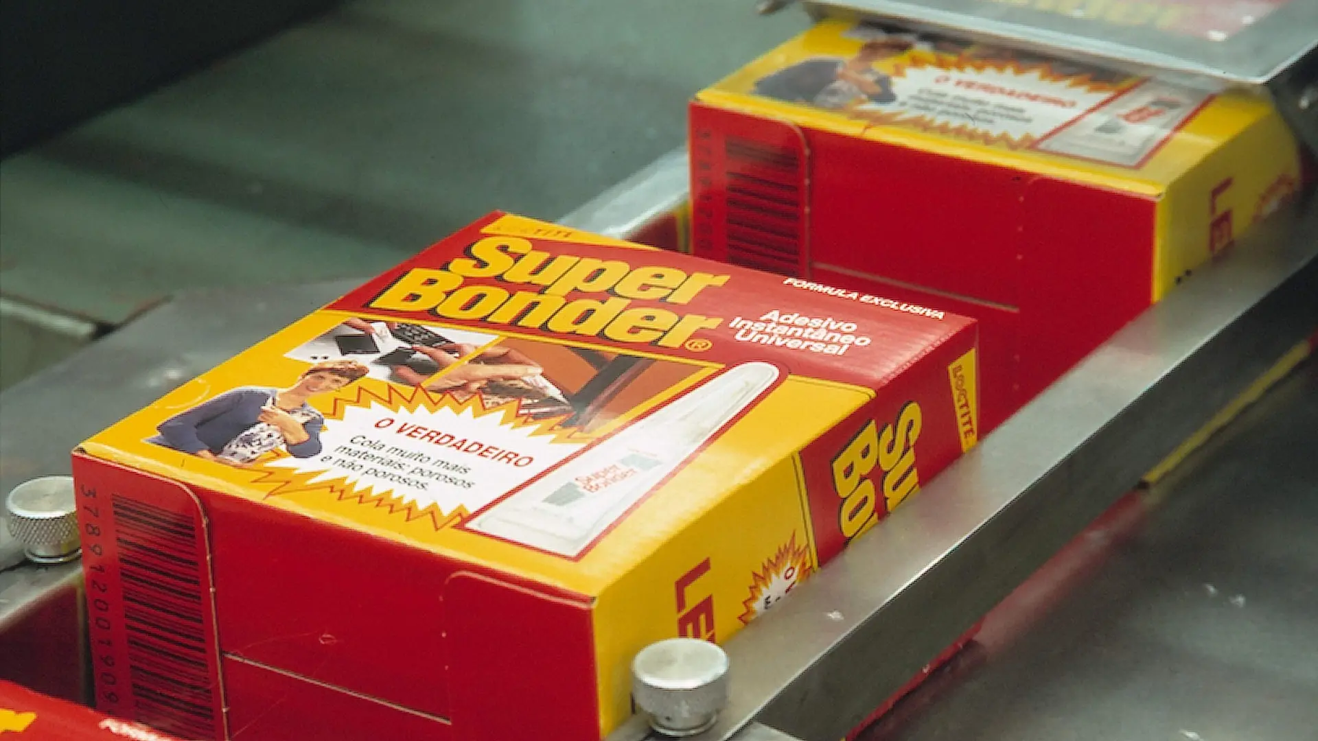 Caixas do adesivo Loctite Super Bonder em esteira de produção. As embalagens são predominantemente vermelhas e amarelas, com o nome 'Super Bonder' em destaque na parte superior e a frase 'O verdadeiro' acompanhada de imagens ilustrativas mostrando aplicações do produto.