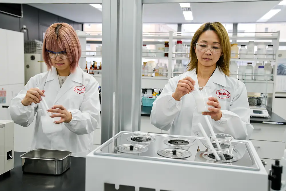 Duas pessoas usando jalecos brancos da Henkel trabalham em um laboratório moderno. Estão manuseando instrumentos científicos e recipientes em uma bancada, com vários equipamentos de laboratório e prateleiras com materiais visíveis ao fundo.