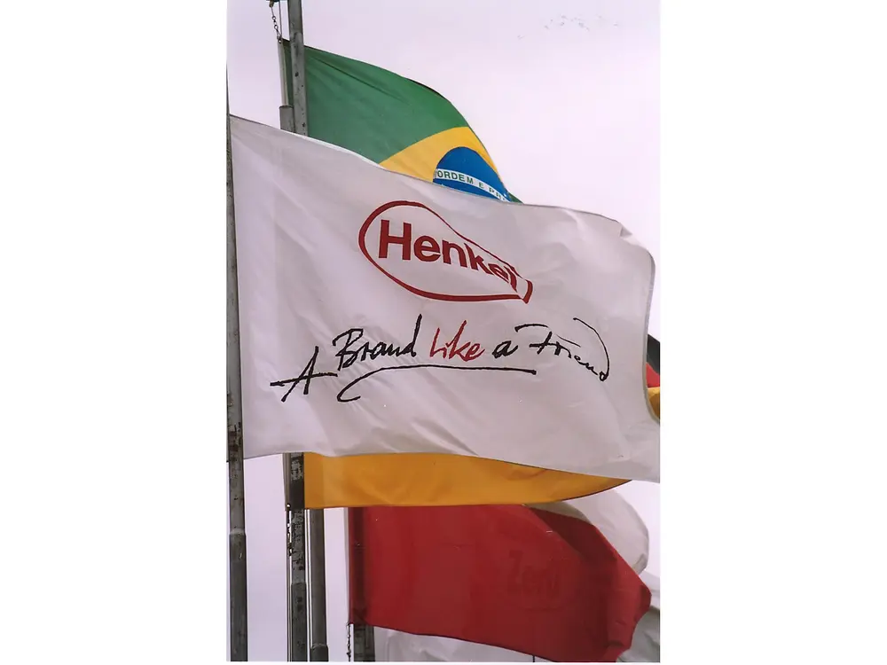 Em destaque na foto, há três bandeiras tremulando. À frente, uma branca com o logo da Henkel. Ao fundo, uma do Brasil e outra da Alemanha.