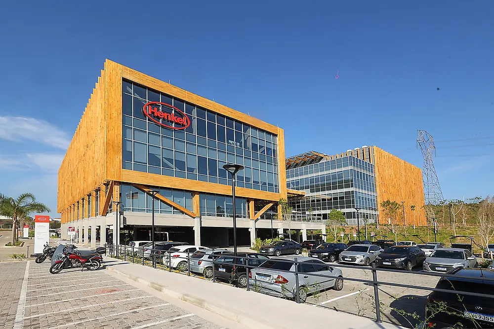 Foto panorâmica do prédio administrativo da Henkel Brasil, que traz o logo da empresa, e do Inspiration Center Jundiaí, em um dia ensolarado. Ambos são estruturados com madeira engenheirada, sendo o principal destaque arquitetônico.