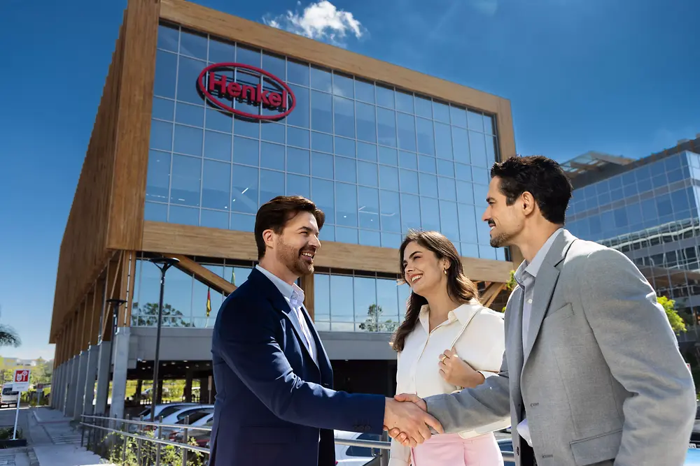 Três pessoas em trajes profissionais conversam e apertam as mãos do lado de fora do Inspiration Center Jundiaí, um moderno edifício de vidro com estrutura de madeira e a placa “Henkel” sob um céu azul claro.