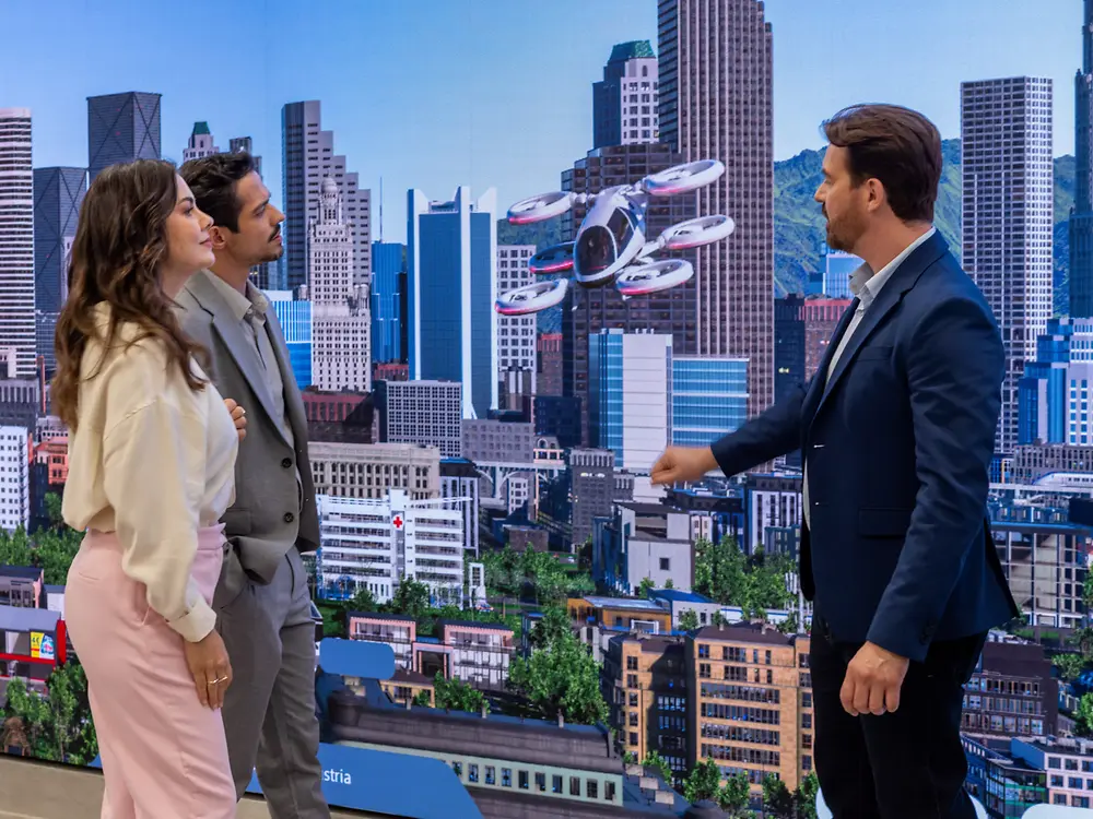 Três pessoas estão dentro da Sala Infinita do Inspiration Center Jundiaí, diante de uma grande tela que exibe uma paisagem urbana futurista com veículos voadores. Elas parecem envolvidas e interessadas.