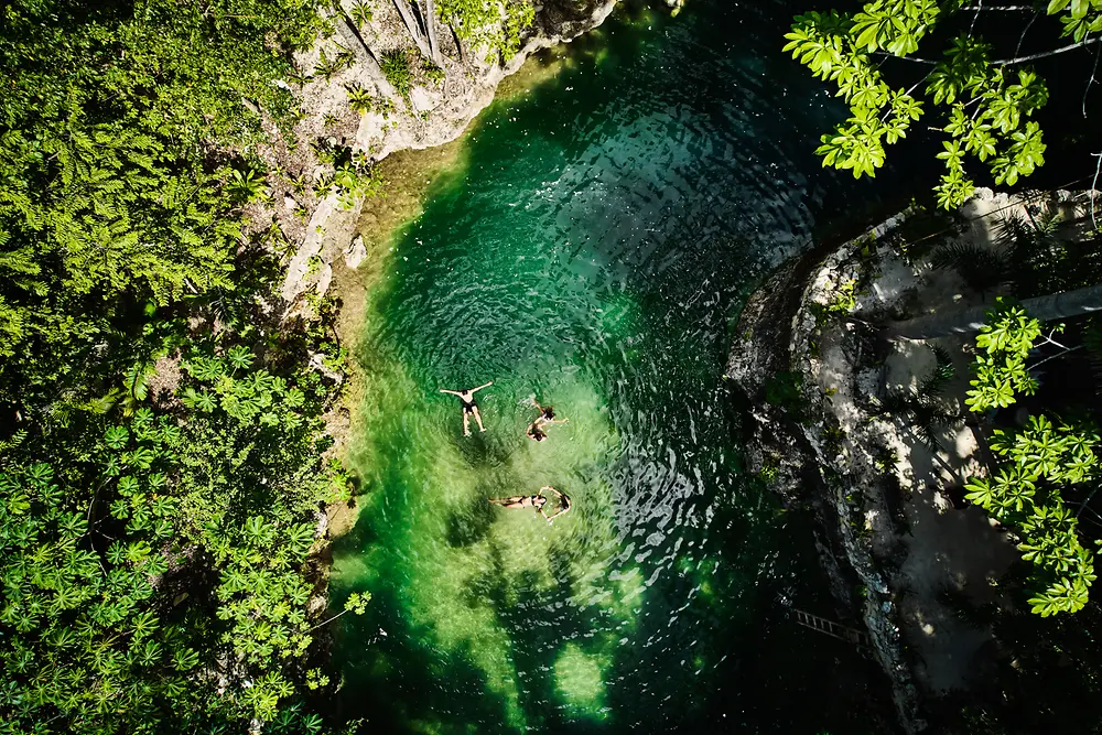 Vista aérea de uma pequena piscina natural cercada por densa vegetação verde, com três pessoas flutuando na água clara, de tom esmeralda.