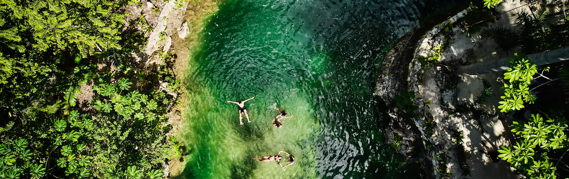 Vista aérea de uma pequena piscina natural cercada por densa vegetação verde, com três pessoas flutuando na água clara, de tom esmeralda.