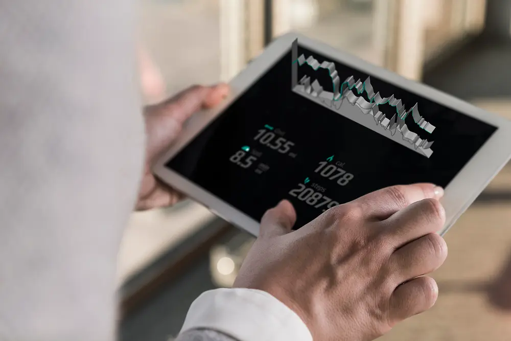 Uma pessoa segurando um tablet que exibe gráficos digitais e dados numéricos, visto por cima do ombro em um ambiente interno bem iluminado.