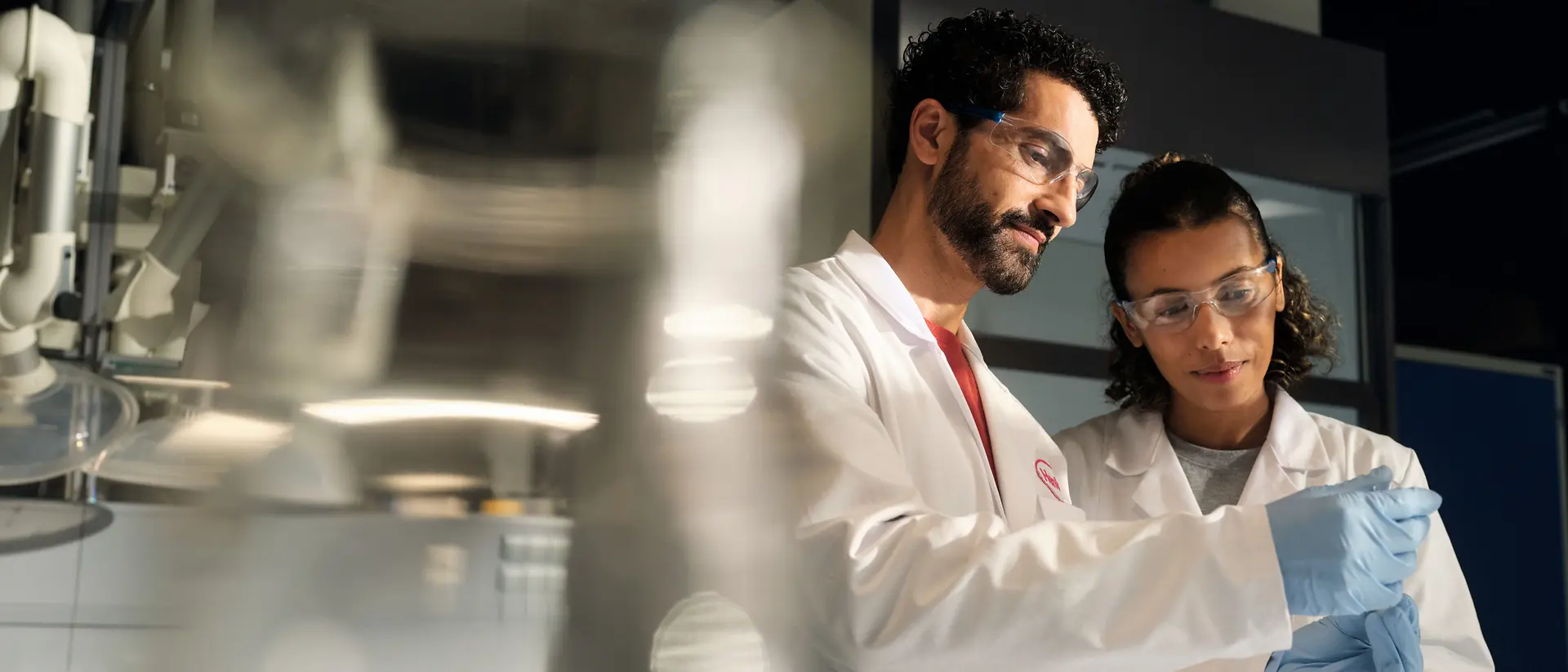Dois cientistas usando jalecos brancos, óculos de proteção e luvas analisam juntos um pequeno recipiente com amostra em um ambiente de laboratório.