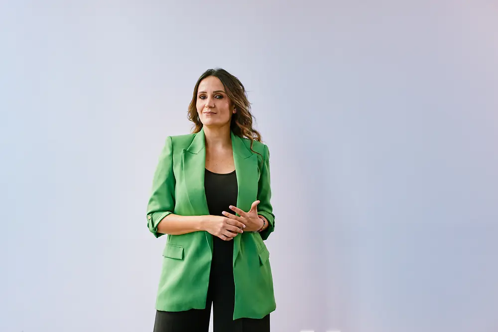 Profissional experiente da Henkel em pé, com postura confiante, diante de uma parede branca em um ambiente moderno, vestindo um blazer verde.